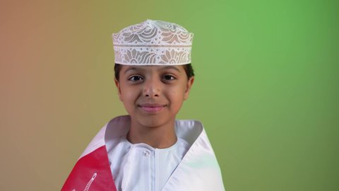 A close-up of an Omani Gulf Arab boy wearing a dishdasha and kummah, holding an Omani flag on his shoulder, looking at the camera with gestures of joy and happiness. Commemorating Omani National Day. The concept of patriotism and belonging to one's homeland, pride in Omani symbols. Omani