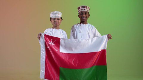 مفهوم حب الوطن والإنتماء إليه ، صديقان عربيان خليجيان عمانيان يرتديان الدشداشة والكمة ، يحملان علم لسلطنة عمان ينظران الى الكاميرا بايماءات الفرح والسرور ، إحياء ذكرى اليوم الوطني العماني، الاعتزاز والفخر بالرموز العمانية