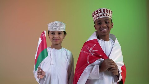 صديقان عربيان خليجيان عمانيان يرتديان الدشداشة والكمة ، يحملان علم لسلطنة عمان ينظران الى الكاميرا بايماءات الفرح والسرور ، إحياء ذكرى اليوم الوطني العماني، الاعتزاز والفخر بالرموز العمانية،  مفهوم حب الوطن والإنتماء إليه