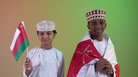 إحياء ذكرى اليوم الوطني العماني، الاعتزاز والفخر بالرموز العمانية،  مفهوم حب الوطن والإنتماء إليه، صديقان عربيان خليجيان عمانيان يرتديان الدشداشة والكمة ، يحملان علم لسلطنة عمان ينظر الى الكاميرا بايماءات الفرح والسرور