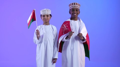 The concept of patriotism and belonging, pride in Omani symbols, commemorating Omani National Day. Two Gulf Arab Omani friends wearing dishdashas and kummah, holding the flag of the Sultanate of Oman, looking at the camera with gestures of joy and happiness.