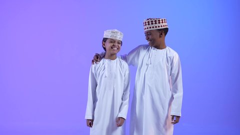 Facial and hand gestures that indicate love and security, a hug and a gathering of friends, two Arab Gulf Omani friends wearing a dishdasha and a kummah, one of them placing his hand on a friend's shoulder, love and support between friends.