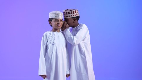 كتمان وحفظ الاسرار، صديقان عربيان خليجيان عمانيان يرتديان الدشداشة والكمة ، صديق يقوم بالهمس في اذن صديقه، التحدث بصوت منخفض، ايماءات وجه تدل على السعادة