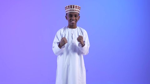 Signs and expressions of joy and strength, admiration, satisfaction, and happiness, achieving goals and accomplishing tasks. A smiling Omani Gulf Arab boy wearing a dishdasha and a kummah raising his fists in gestures of victory, triumph, and gain.