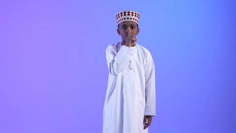 التوقف عن الكلام و الأمر بالصمت و الاستماع، ولد عربي خليجي عماني مبتسم يرتدي الدشداشة والكمة، يرفع اصبعه الى فمه, إيماءات يدين تدل على طلب الهدوء