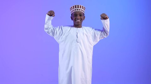 Admiration, satisfaction, and a feeling of happiness, achieving goals and completing tasks. A smiling Omani Gulf Arab boy wearing a dishdasha and a kummah raising his hands in gestures of victory, triumph, and gain. Signs and expressions of joy and strength.