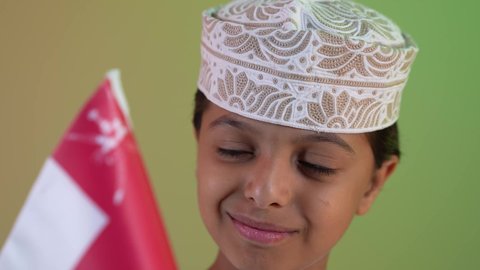 مفهوم حب الوطن والإنتماء إليه، الاعتزاز والفخر بالرموز العمانية، إحياء ذكرى اليوم الوطني العماني، تصوير مقرب لولد عربي خليجي عماني يرتدي الدشداشة والكمه يمسك علم لسلطنة عمان ينظر الى العلم بايماءات الفرح والسرور