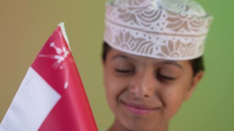 الاعتزاز والفخر بالرموز العمانية،مفهوم حب الوطن والإنتماء إليه، إحياء ذكرى اليوم الوطني العماني، تصوير مقرب لولد عربي خليجي عماني يرتدي الدشداشة والكمه يمسك علم لسلطنة عمان ينظر الى العلم بايماءات الفرح والسرور