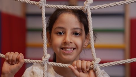 Expressions of joy and pleasure while playing, promoting physical activity and balance, practicing motor activities for children, a Saudi Arabian Gulf girl wearing casual clothes, holding ropes on a climbing net inside a children's gym, developing physical skills.