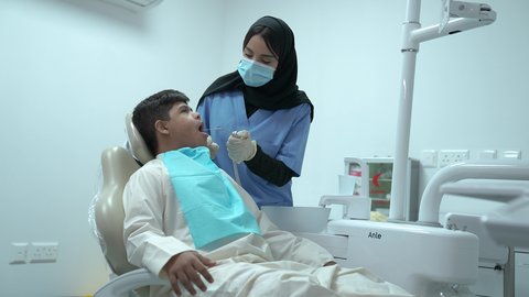 المعدات اللازمة لإجراء الفحوصات، طبيبة عربية خليجية سعودية ترتدي الكمامة والقفازات الطبية تقوم بفحص أسنان المريض ، ولد عربي خليجي سعودي مصاب بمتلازمة داون يرتدي الزي التقليدي ، يجلس في العيادة السنية ويفتح فمه لتشخيص المرض ،معالجة النخر والتسوس