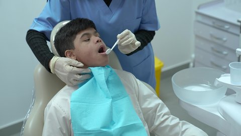 ولد عربي خليجي سعودي مصاب بمتلازمة داون يرتدي الزي التقليدي ، يجلس في العيادة السنية ويفتح فمه لتشخيص المرض ،معالجة النخر والتسوس,  الادوات اللازمة لإجراء الفحوصات، طبيبة عربية خليجية سعودية ترتدي الكمامة والقفازات الطبية تقوم بفحص أسنان المريض