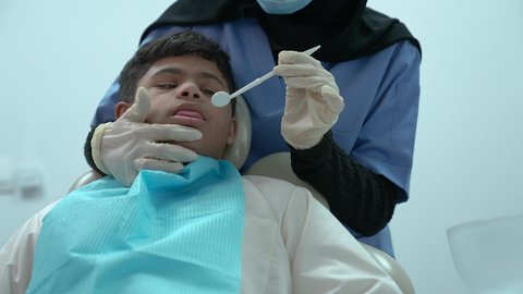 معالجة النخر والتسوس,  الادوات اللازمة لإجراء الفحوصات، ولد عربي خليجي سعودي مصاب بمتلازمة داون يرتدي الزي التقليدي ، يجلس في العيادة السنية ويفتح فمه لتشخيص المرض ، طبيبة عربية خليجية سعودية ترتدي الكمامة والقفازات الطبية تقوم بفحص أسنان المريض