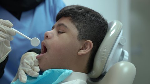 الادوات اللازمة لإجراء الفحوصات، معالجة النخر والتسوس, ولد عربي خليجي سعودي مصاب بمتلازمة داون يرتدي الزي التقليدي ، يجلس في العيادة السنية ويفتح فمه لتشخيص المرض ، طبيبة عربية خليجية سعودية ترتدي الكمامة والقفازات الطبية تقوم بفحص أسنان المريض