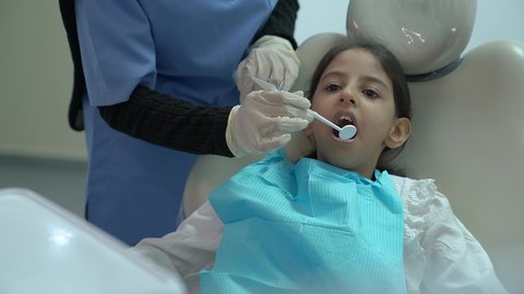 بنت عربية خليجية سعودية ترتدي الزي الكاجوال ، تجلس في العيادة السنية وتفتح فمها لتشخيص المرض ،طبيبة عربية خليجية سعودية ترتدي الكمامة والقفازات الطبية تقوم بفحص أسنان المريض ،  معالجة النخر والتسوس،  المعدات اللازمة لإجراء الفحوصات