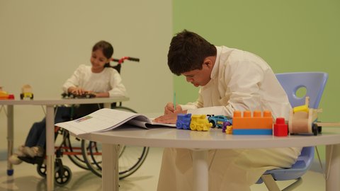 تنمية المهارات الفنية في الروضات السعودية ،إيماءات التركيز والاستمتاع بالرسم و التلوين، مفهوم الاهتمام بالهوايات, ولد عربي خليجي سعودي مصاب بمتلازمة داون يرتدي الزي التقليدي يقوم بالرسم والتلوين بالألوان ، قضاء اوقات ممتعه