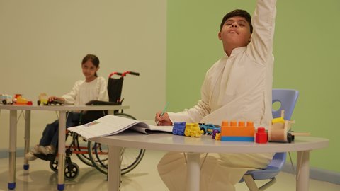 إيماءات التركيز والاستمتاع بالرسم و التلوين، مفهوم الاهتمام بالهوايات,تنمية المهارات الفنية في الروضات السعودية ، ولد عربي خليجي سعودي مصاب بمتلازمة داون يرتدي الزي التقليدي يقوم بالرسم والتلوين بالألوان ، قضاء اوقات ممتعه