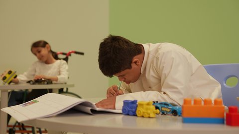 The concept of interest in hobbies, gestures indicating focus and enjoyment in drawing and coloring, developing artistic skills in Saudi kindergartens, a Saudi Arabian boy with Down syndrome wearing traditional clothing, drawing and coloring, having a good time.