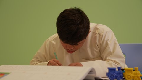 The concept of interest in hobbies, developing artistic skills in Saudi kindergartens, gestures of concentration and enjoyment in drawing and coloring, a Saudi Arabian boy with Down syndrome wearing traditional clothing, drawing and coloring, having a good time.