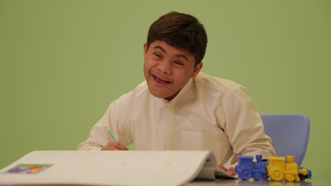 Developing artistic skills in Saudi kindergartens, gestures of concentration and enjoyment of drawing and coloring, the concept of interest in hobbies, a Saudi Arabian Gulf boy with Down syndrome wearing traditional clothing, drawing and coloring, having a good time.