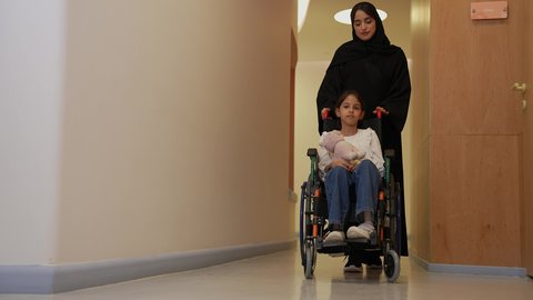 بيئة طبية آمنة ، مفهوم الإنسانية والرحمة، الرعاية الصحية والدعم النفسي للمرضى وذوي الاحتياجات الخاصة، امرأة عربية خليجية سعودية ترتدي العباءة السوداء تقوم بدفع فتاة صغيرة مبتسمة تجلس على كرسي متحرك  في المستشفى ، ايماءات العناية والاهتمام
