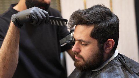 Taking care of one's appearance and physical hygiene, providing personal care services, practicing the barbering profession, a close-up photo from the side of a Saudi Arabian Gulf barber shaving a customer's hair using a razor