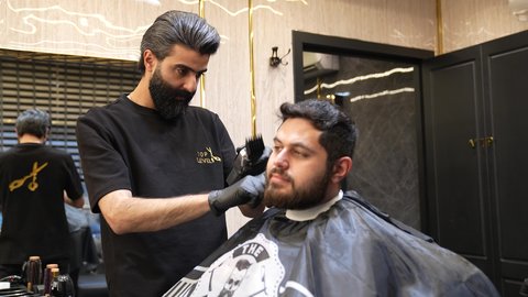 الاهتمام بالشكل الخارجي، الممارسة مهنه الحلاقة، تقديم خدمات العناية الشخصية, حلاق عربي خليجي سعودي يرتدي يونيفورم الصالون يقوم بقص شعر الزبون باستخدام ماكينه الحلاقة