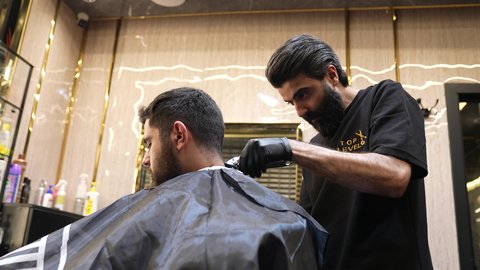زيارة روتينية لصالون الحلاقة، الممارسة مهنه الحلاقة، تقديم خدمات العناية الشخصية, تصويرمن  الجانب لحلاق عربي خليجي سعودي يرتدي يونيفورم الصالون يقوم بقص شعر الزبون باستخدام ماكينه الحلاقة