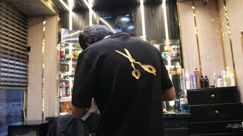 جلسة حلاقة شعر وذقن، الممارسة مهنه الحلاقة، تقديم خدمات العناية الشخصية, تصويرمن الخلف لحلاق عربي خليجي سعودي يرتدي يونيفورم الصالون يقوم بقص شعر الزبون باستخدام ماكينه الحلاقة