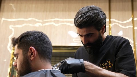 خدمات العناية الرجالية، الممارسة مهنه الحلاقة، تقديم خدمات العناية الشخصية, تصويرمقرب لحلاق عربي خليجي سعودي يرتدي يونيفورم الصالون يقوم بقص شعر الزبون باستخدام ماكينه الحلاقة