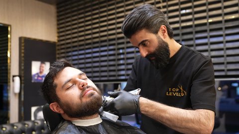 الممارسة مهنه الحلاقة، المحافظة على الاناقة والنظافة الشخصية، صالون تجميلي رجالي لتصفيف الشعر، تقديم خدمات العناية الشخصية, تصوير لحلاق عربي خليجي سعودي يرتدي يونيفورم الصالون يقوم بحلاقة ذقن الزبون
