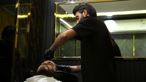 العناية والإهتمام بشعر صحي ونظيف، ممارسة مهنة الحلاقة, تقديم خدمات العناية الشخصية, الاهتمام بالشعر و النظافة البدنية، تصوير لحلاق عربي خليجي سعودي يرتدي يونيفورم الصالون يقوم بغسيل شعر الزبون