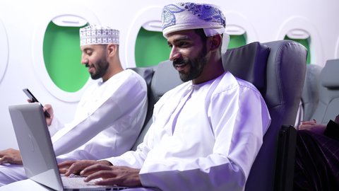 مفهوم ممارسه الانشطه والعمل عن بعد داخل الطائره, تصوير مقرب من الجانب لرجل عربي خليجي عماني يرتدي الدشداشه والكمه يجلس على كرسي داخل الطائره الخليجيه يقوم بالعمل على جهاز الحاسوب المحمول, بجانبه شاب يستخدم الجوال