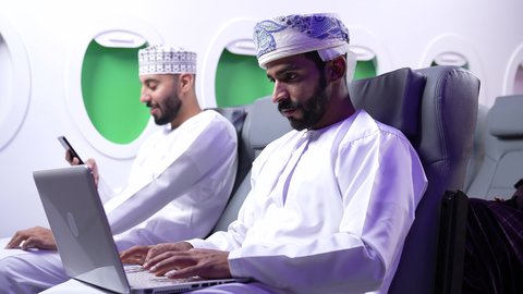 Using modern technology, the concept of practicing activities and working remotely inside an airplane. A close-up side view of an Arab Gulf Omani man wearing a dishdasha and a keffiyeh sitting on a chair inside a Gulf airplane working on a laptop. Next to him is a young man using a mobile phone.