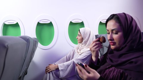 الاهتمام والعنايه بالمظهر الخارجي, التسويق لمستحضرات التجميل, تصوير مقرب من الجانب لامرأه عربيه خليجيه عمانيه ترتدي العباءة والحجاب تضع المكياج على وجهها داخل الطائره باستعمال فرشاة أحمر الخدود ، بجانب سيدة سعودية تسافر لقضاء اجازة العطله