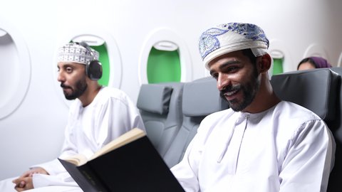 ممارسه الهوايات المفضله ،  الاستمتاع بقرائه القصص والروايات, تصوير مقرب من الجانب لرجل عربي خليجي عماني يرتدي الزي العماني يجلس على كراسي الطائره في رحله جويه يقرأ الرجل كتابا ، بجانبه شاب يرتدي سماعات الراس، الترفيه والتسليه وقضاء الاوقات داخل الطائره