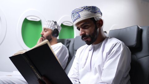 الاستمتاع بقرائه القصص والروايات, ممارسه الهوايات المفضله ، تصوير مقرب من الجانب لرجل عربي خليجي عماني يرتدي الزي العماني يجلس على كراسي الطائره في رحله جويه يقرأ الرجل كتابا ، بجانبه شاب يرتدي سماعات الراس، الترفيه والتسليه وقضاء الاوقات داخل الطائره