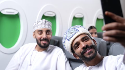 التقاط صور السيلفي ، الترفيه والتسليه وقضاء الاوقات داخل الطائره ، دمج التقنيات فى الحياه اليوميه ، تصوير مقرب لصديقان عربيان خليجيان عمانيان يجلسان على كراسي الطائره في رحله جويه