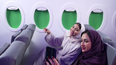Entertainment, amusement, and spending time on board an airplane. Integrating technology into daily life. A Saudi woman takes selfies. A close-up of two Arab Gulf and Omani friends sitting on airplane seats on a flight.
