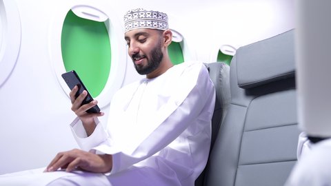Checking on family and friends, browsing social media, a close-up of a Gulf Arab Omani man wearing a dishdasha and a keffiyeh sitting on a seat inside a Gulf plane using a mobile phone, integrating modern technology into daily life.
