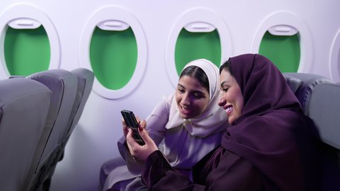 دمج التقنيات فى الحياه اليوميه ،الترفيه والتسليه وقضاء الاوقات داخل الطائره ، سيدة سعودية تلتقط صور السيلفي ،  تصوير مقرب لصديقتان عربيتان خليجيتان عمانيتان تجلسان على كراسي الطائره في رحله جويه