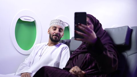 دمج التقنيات فى الحياه اليوميه ،الترفيه والتسليه وقضاء الاوقات داخل الطائره ، التقاط صور السيلفي ،  تصوير مقرب لرجل وامرأه عربييان خليجييان عمانييان يجلسان على كراسي الطائره في رحله جويه