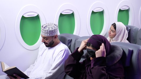 الترفيه والتسليه وقضاء الاوقات الممتعه داخل الطائره , تصوير مقرب من الجانب لامرأه عربيه خليجيه عمانيه ترتدي العباءة والحجاب تجلس على كرسي داخل الطائره الخليجيه ، تضع منامه العين ومخده الرأس, بجانبها شاب عماني يقرأ كتاباً