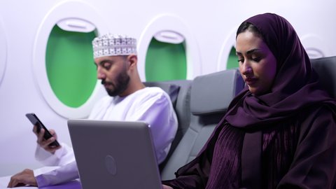 استخدام التقنيات الحديثة ، مفهوم ممارسه الانشطه والعمل عن بعد داخل الطائره, تصوير مقرب من الجانب لامرأه عربيه خليجيه عمانيه ترتدي العباءة والحجاب تجلس على كرسي داخل الطائره الخليجيه تقوم بالعمل على جهاز الحاسوب المحمول, بجانبها شاب عماني يستخدم الجوال