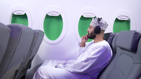 Listening to music, having a good time on an airplane, having fun, entertaining, and integrating while traveling. An Arab Gulf Omani man wearing a dishdasha and a keffiyeh sits on a seat inside a Gulf airplane on a flight.