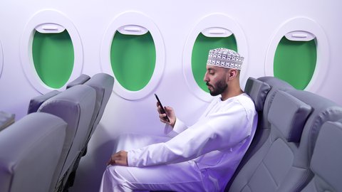 Integrating modern technologies into daily life, browsing social media, checking on family and friends, a close-up of a Gulf Arab Omani man wearing a dishdasha and a keffiyeh sitting on a seat inside a Gulf airplane using a mobile phone.