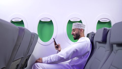 تصفح مواقع التواصل الاجتماعي , الاطمئنان على الاهل والاصدقاء ، تصوير مقرب من الجانب لرجل عربي خليجي عماني يرتدي الدشداشه والكمه يجلس على كرسي داخل الطائره الخليجيه يقوم باستخدام الهاتف المحمول, دمج التقنيات الحديثة فى الحياة اليومية