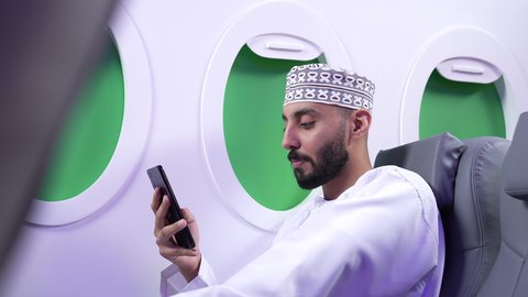 الاطمئنان على الاهل والاصدقاء ، تصفح مواقع التواصل الاجتماعي , تصوير مقرب من الجانب لرجل عربي خليجي عماني يرتدي الدشداشه والكمه يجلس على كرسي داخل الطائره الخليجيه يقوم باستخدام الهاتف المحمول, دمج التقنيات الحديثة فى الحياة اليومية