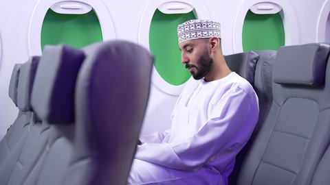 الترفيه والتسليه وقضاء الاوقات داخل الطائره، ممارسه الهوايات المفضله ، الاستمتاع بقرائه القصص والروايات,تصوير مقرب من الجانب لرجل عربي خليجي عماني يرتدي الزي العماني يجلس على كراسي الطائره في رحله جويه يقرأ الرجل كتابا