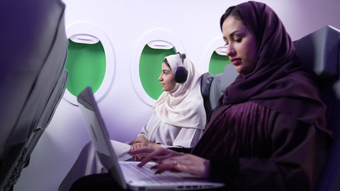 قضاء الاوقات السعيده داخل الطائره, استخدام التكنولوجيا والتقنيات الحديثه ، الاستماع الى الموسيقي ، التسليه والترفيه والاندماج مع العائله، امرأه عربيه خليجيه عمانيه ترتدي العباءة والحجاب تستخدم جهاز اللابتوب ، سيدة سعودية تجلس على كرسي داخل الطائره في رحله جويه وترتدي سماعات الرأس