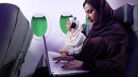 الاستماع الى الموسيقي ، استخدام التكنولوجيا والتقنيات الحديثه ، قضاء الاوقات السعيده داخل الطائره, التسليه والترفيه والاندماج مع العائله، امرأه عربيه خليجيه عمانيه ترتدي العباءة والحجاب تستخدم جهاز اللابتوب ، سيدة سعودية تجلس على كرسي داخل الطائره في رحله جويه وترتدي سماعات الرأس